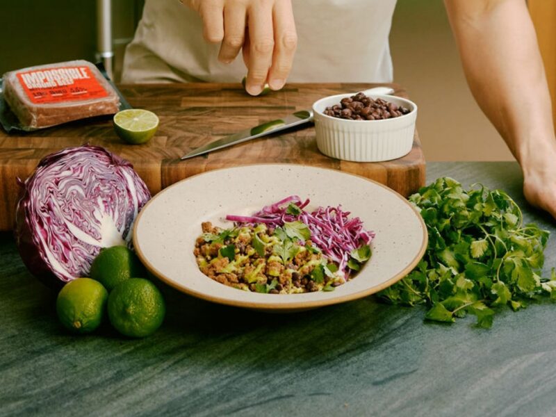 A man standing in a kitchen prepares a Purple Carrot plant based meal. Purple cabbage, cilantro, and Impossible meat can be seen around a white bowl.