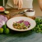 A man standing in a kitchen prepares a Purple Carrot plant based meal. Purple cabbage, cilantro, and Impossible meat can be seen around a white bowl.
