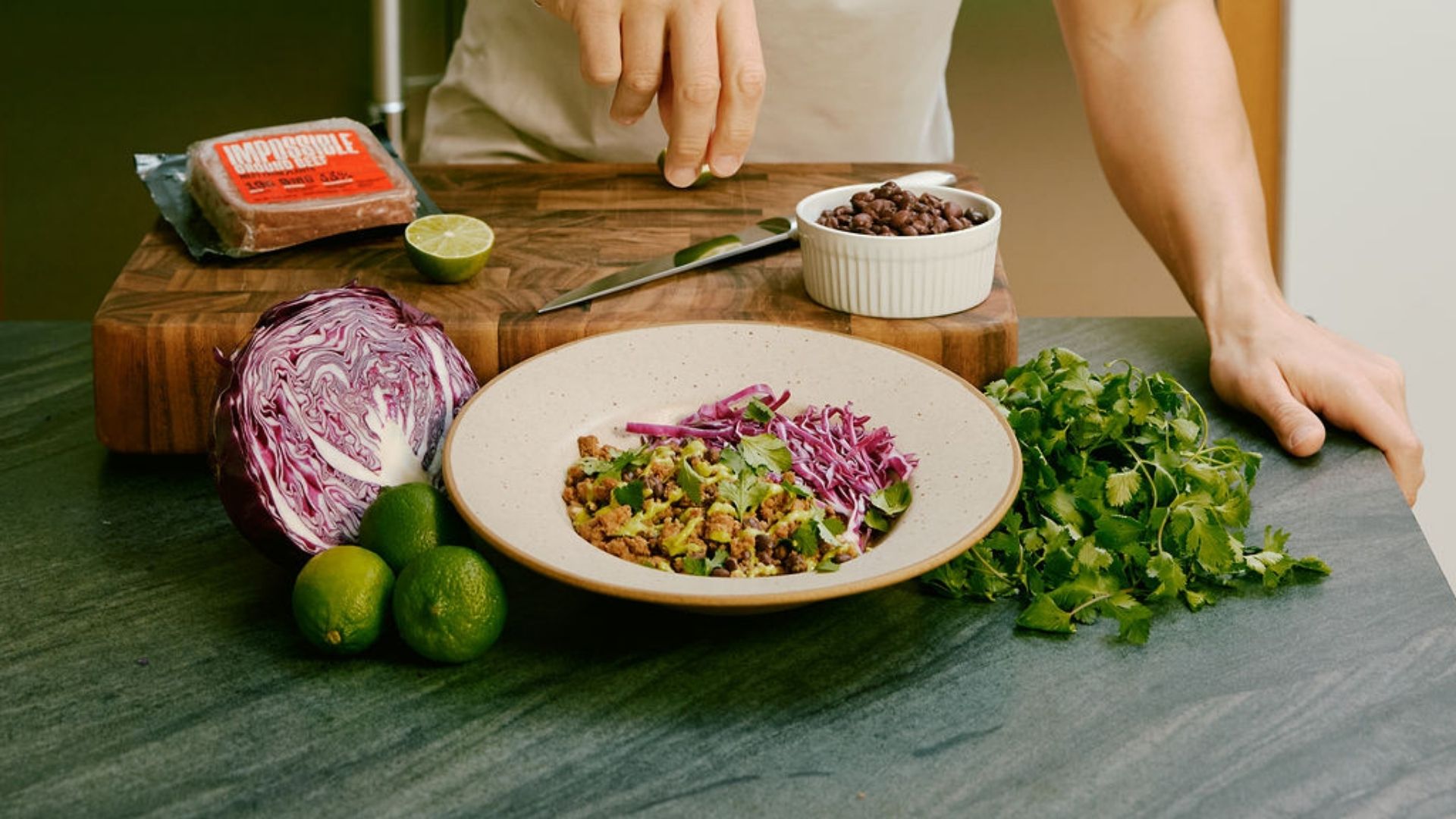 A man standing in a kitchen prepares a Purple Carrot plant based meal. Purple cabbage, cilantro, and Impossible meat can be seen around a white bowl.