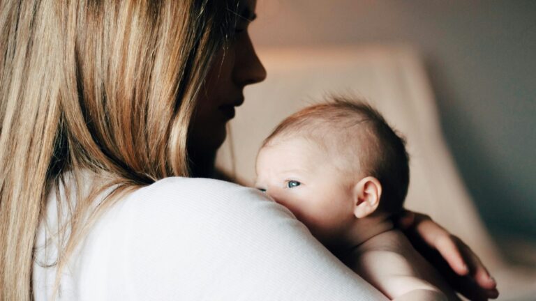 A postpartum parent with long blonde hair and wearing a white short holds a newborn baby