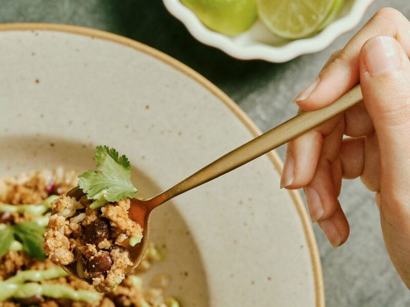 A white hand holds a spoon digging into a delicious vegan meal from Purple Carrot. The meal primarily features beans, a great source of plant based protein.