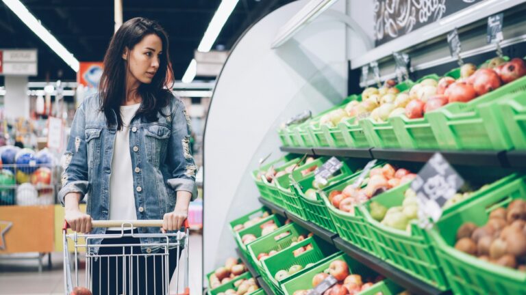 An Asian woman pushes a grocery cart up the produce aisle of a grocery store.