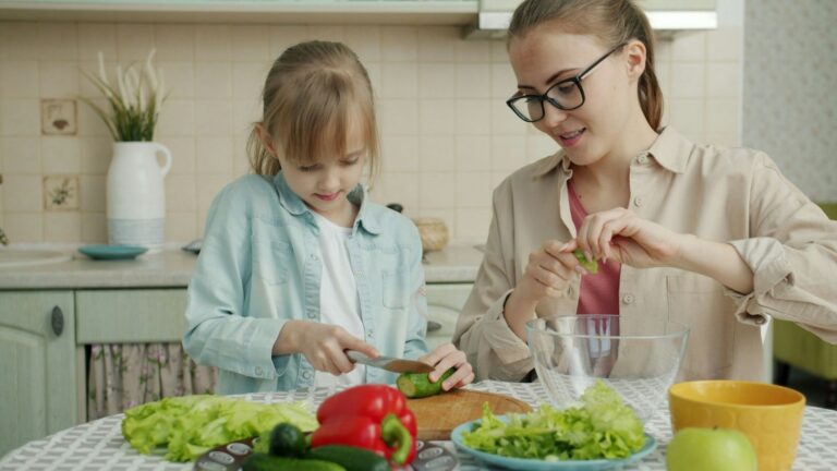 A mother and daughter sit in a kitchen. The daughter is practicing cutting vegetables with a knife. Lettuce and red bell peppers are in the foreground.