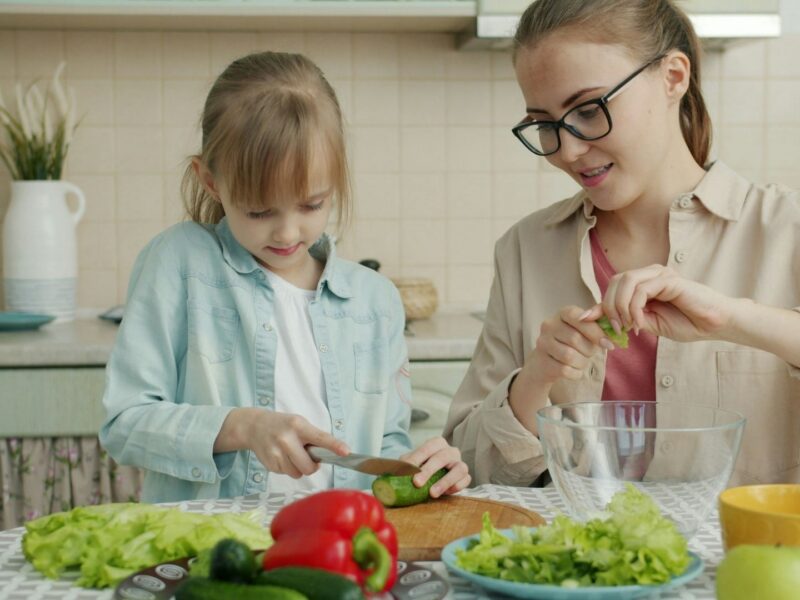 A mother and daughter sit in a kitchen. The daughter is practicing cutting vegetables with a knife. Lettuce and red bell peppers are in the foreground.