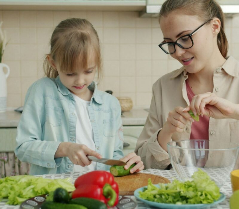 A mother and daughter sit in a kitchen. The daughter is practicing cutting vegetables with a knife. Lettuce and red bell peppers are in the foreground.