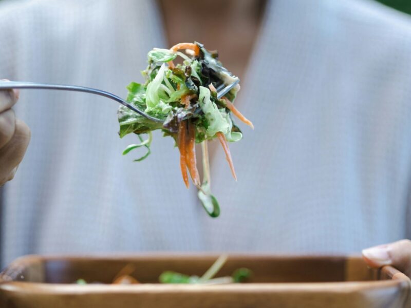 A person wearing a white shirt eats a heart-healthy salad. The salad is in a wooden bowl and is prominent in the foreground of the photo.