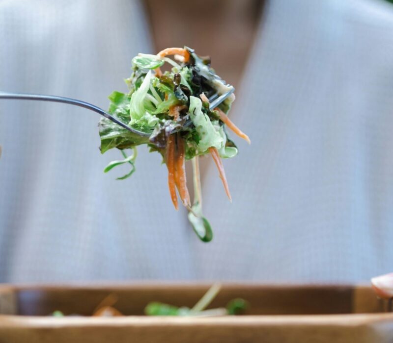 A person wearing a white shirt eats a heart-healthy salad. The salad is in a wooden bowl and is prominent in the foreground of the photo.