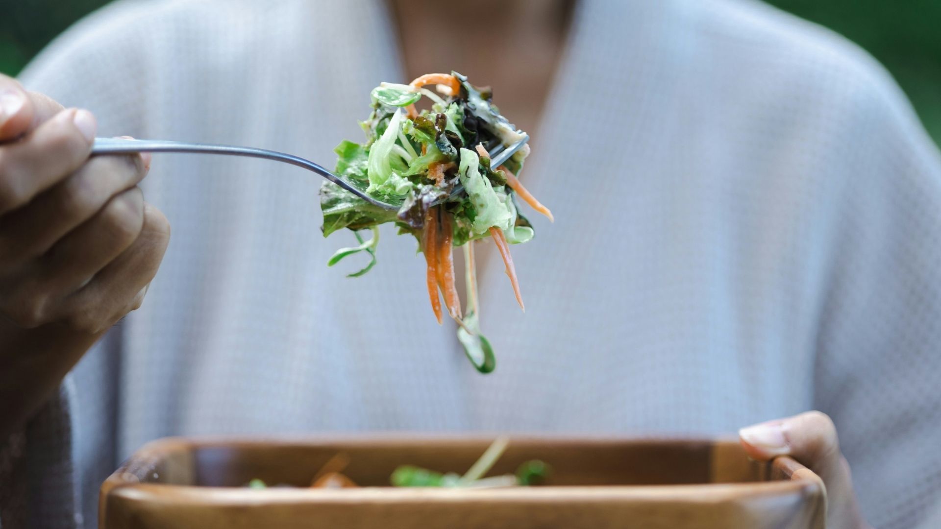 A person wearing a white shirt eats a heart-healthy salad. The salad is in a wooden bowl and is prominent in the foreground of the photo.