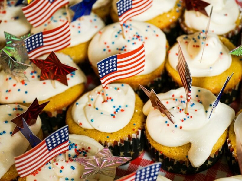 A close up of a cupcake tray featuring vegan cupcakes. Each cupcake has an American flag toothpick for Memorial Day.