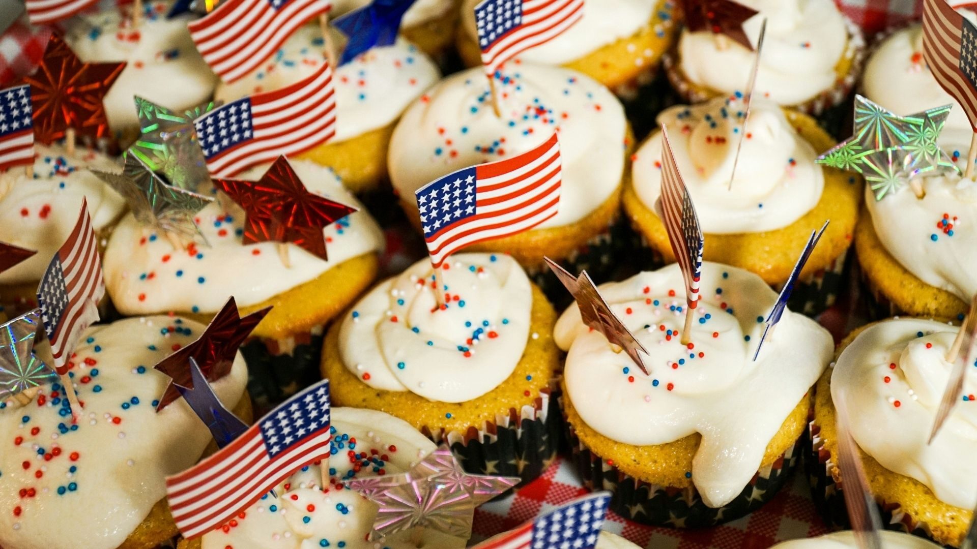 A close up of a cupcake tray featuring vegan cupcakes. Each cupcake has an American flag toothpick for Memorial Day.
