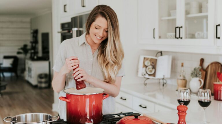 A blonde woman stands in a kitchen. She is holding a pepper grinder over a red ceramic pot as she cooks seitan.