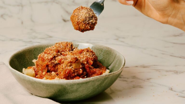 Plant-based meatballs from Purple Carrot are piled in a green bowl. A fork holds a singular meatball above the bowl.
