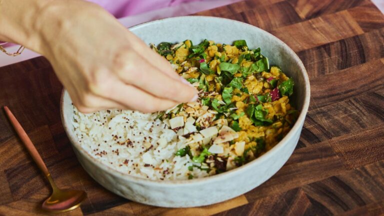 A Purple Carrot meal featuring plant-based substitutes sits on a wooden countertop. A hand sprinkles sesame seeds on top of the meal.